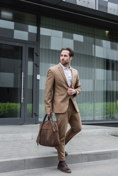 full length of bearded businessman in suit holding leather bag on urban street - Zdjęcie, obraz
