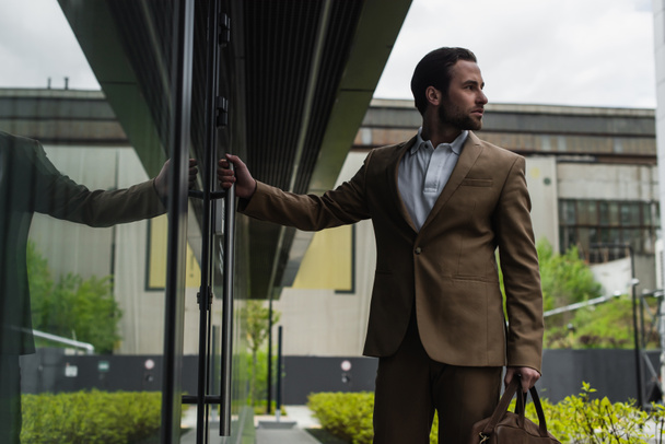 бородатый бизнесмен в формальной одежде держит кожаную сумку возле здания снаружи - Фото, изображение