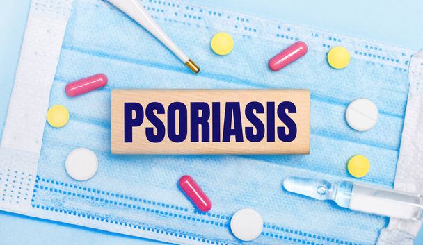 On a light blue disposable face mask there are tablets, a thermometer, an ampoule and a wooden block with the text PSORIASIS. Medical concept - Фото, изображение