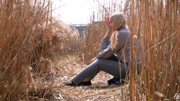 a woman is sitting thoughtfully along the reed field by the river , clear day, blue sky - Imágenes, Vídeo