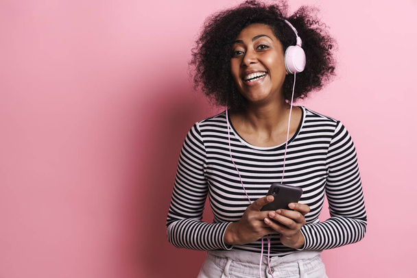 Joyful african american girl listening music with cellphone and headphones isolated over pink background - Foto, Imagem