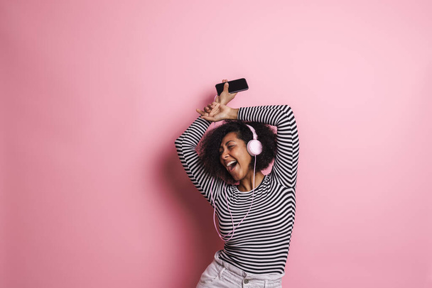 Cheerful young attractive african woman listening to music with headphones isolated over pink background, dancing - Photo, Image