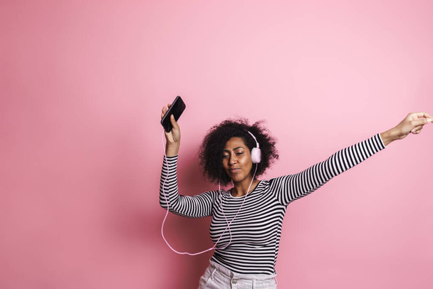 Cheerful young attractive african woman listening to music with headphones isolated over pink background, dancing - Fotografie, Obrázek