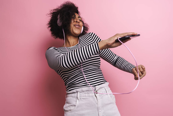 Cheerful young attractive african woman listening to music with headphones isolated over pink background, dancing - Fotografie, Obrázek