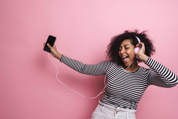 Funny african american girl listening music with cellphone and headphones isolated over pink background - Photo, Image
