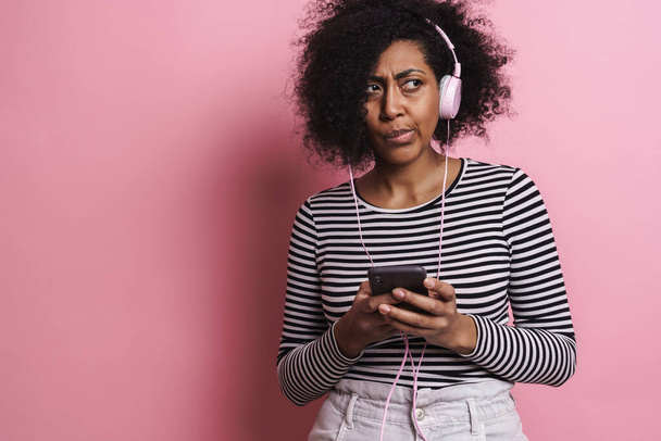 Fille afro-américaine confus écouter de la musique avec téléphone portable et écouteurs isolés sur fond rose - Photo, image