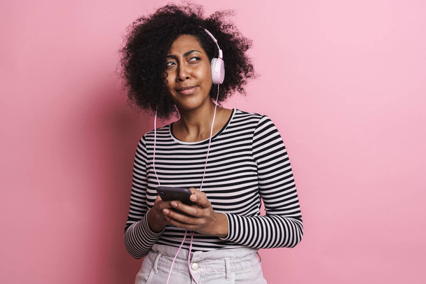 Menina americana africana confusa ouvindo música com celular e fones de ouvido isolados sobre fundo rosa - Foto, Imagem