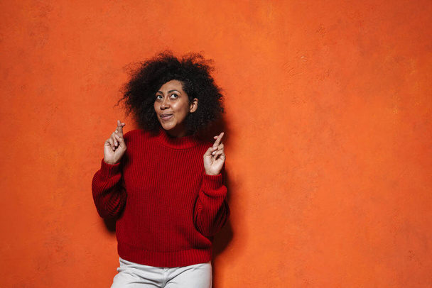 Attractive worried young casual african woman standing isolated over orange background, holding fingers crossed for good luck - Fotografie, Obrázek