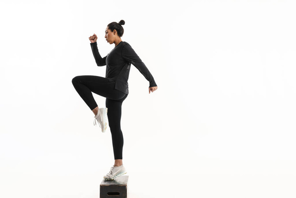 Afro american sportswoman with outstretched hands jumping om wooden fitness block isolated on white - 写真・画像