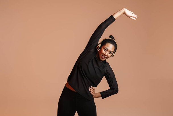 Smiling african american woman in headphones doing exercise while working out isolated over beige background - Foto, Imagem