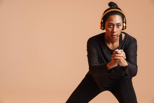 Focused african american woman in headphones doing exercise while working out isolated over beige background - Foto, Imagem