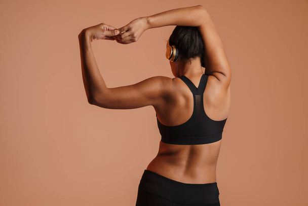 African american woman in headphones doing exercise while working out isolated over beige background - Foto, Imagem
