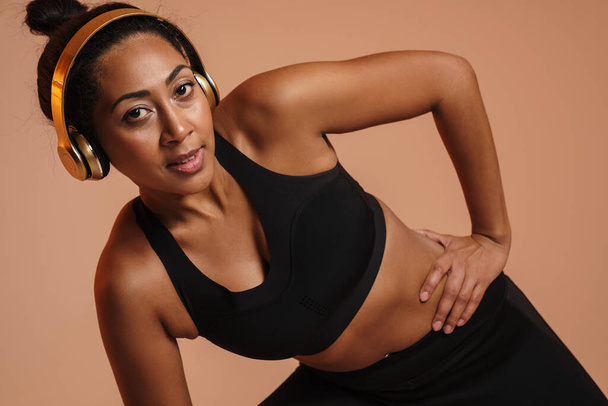 Pleased african american woman in headphones doing exercise while working out isolated over beige background - Photo, image