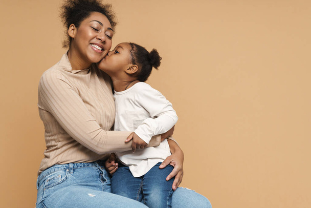 Happy black mother and daughter hugging and kissing isolated over beige background - Фото, изображение