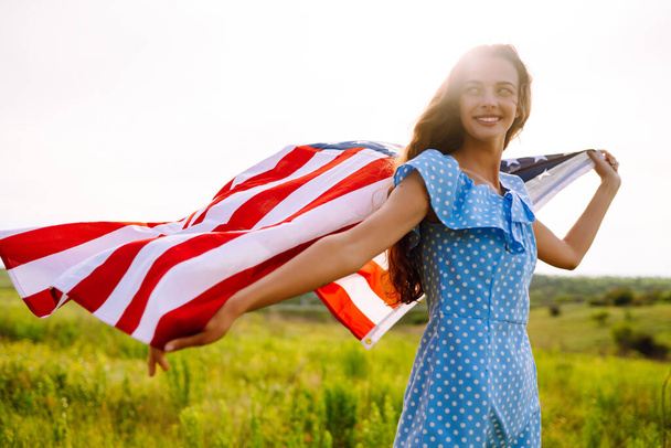 Ευτυχισμένη γυναίκα που ποζάρει με την εθνική σημαία των ΗΠΑ να στέκεται στο ανθισμένο λιβάδι. 4 Ιουλίου. Ημέρα Ανεξαρτησίας. Πατριωτικές διακοπές. - Φωτογραφία, εικόνα