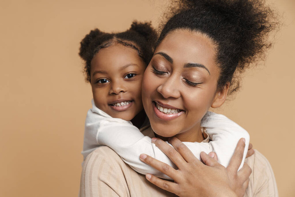 Happy black mother and daughter hugging and smiling isolated over beige background - Фото, изображение