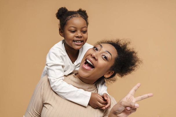 Happy black mother and daughter laughing while showing peace sign isolated over beige background - Фото, изображение