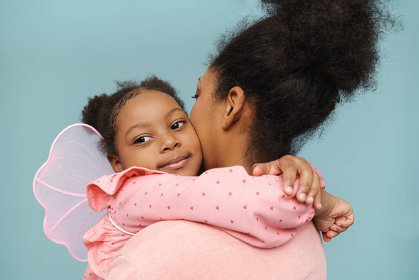 Happy black mother and daughter with butterfly wings hugging and smiling isolated over blue background - Foto, Imagen