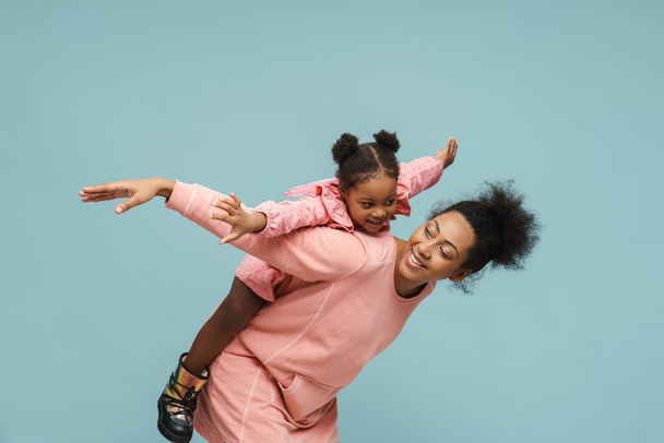 Joyful black mother and daughter with butterfly wings doing piggyback ride isolated over blue background - Фото, зображення