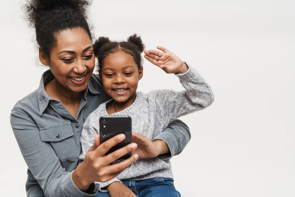 African mother and daughter gesturing while using cellphone isolated over white wall - Foto, afbeelding