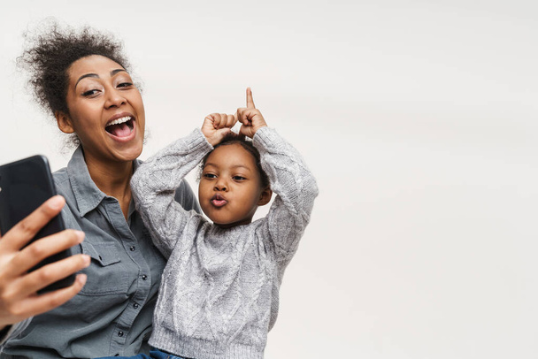 African mother and daughter making fun while using cellphone isolated over white wall - Fotografie, Obrázek