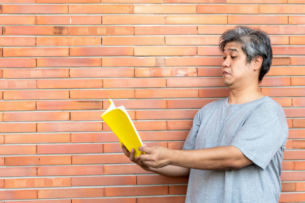 Hombre asiático de mediana edad tiene el pelo gris, está leyendo un libro al mantenerlo alejado de los ojos, porque tiene hipermetropía, con fondo de pared, a la gente y Degeneración de la retina. - Foto, Imagen