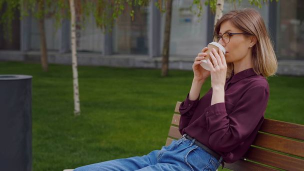 Επιχειρηματίας κάνει ένα διάλειμμα από την εργασία και κάθεται σε ένα παγκάκι έξω από το γραφείο της, ενώ πίνοντας καφέ για να πάει. Φορώντας περιστασιακό στυλ. Ηλιόλουστη μέρα. Πορτρέτο αρκετά επιτυχημένη λευκή κυρία. Βίντεο αργής κίνησης. - Φωτογραφία, εικόνα