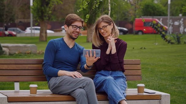 Молодая улыбающаяся пара, проводящая видеозвонок по телефону. Красивая белая женщина и мужчина сидят на открытом пляже кофе-брейк. Видеозвонок по телефону. - Фото, изображение