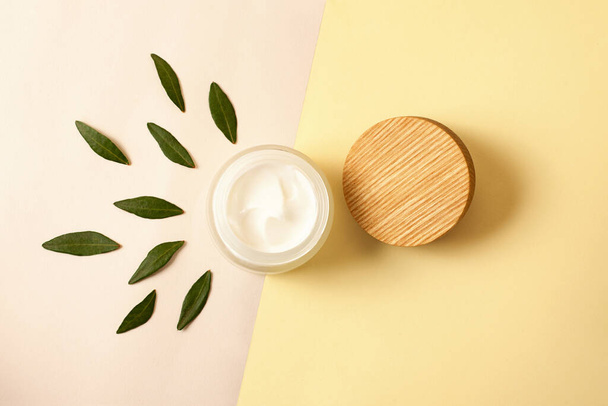 Top view of the zero waste cosmetics container with green leafs around.Open skin care packaging with white cream and wooden cover near it.Concept of organic cosmetic. - Photo, Image
