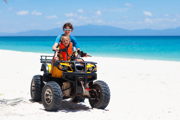 Teenager and his little brother riding quad bike on tropical beach. Active teen age boy on quadricycle. All-terrain vehicle ride. Motor cross sports on ocean sand dune. Kids summer vacation activity. - Photo, Image