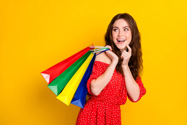 Photo of positive charming girl finger on chin arm hold bags look empty space isolated on yellow color background - Fotoğraf, Görsel