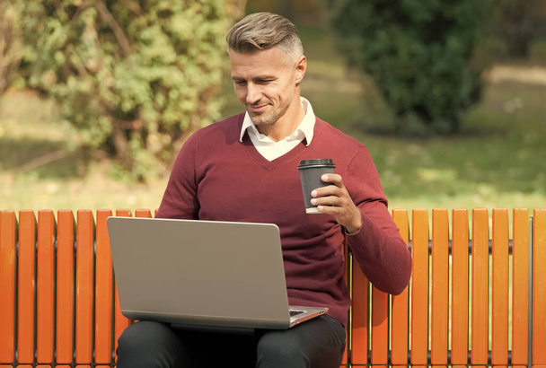 nice start of the day. good morning. mature handsome man work on computer outdoor. well groomed man sit on bench with coffee. confident businessman is online. casual business fashion. agile business - Foto, Imagem