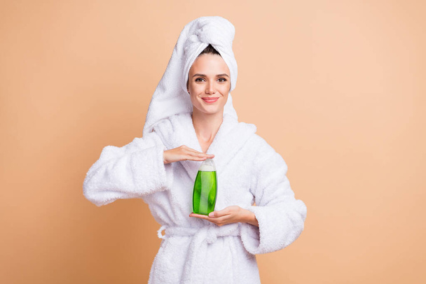 Photo of happy young woman show shampoo bottle wear turban bathroom outfit isolated on beige color background - Valokuva, kuva