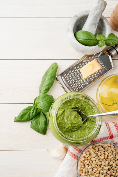 Italian cuisine. Preparing homemade italian pesto sauce. Homemade pesto sause in a glass jar with ingredients, top view flat lay on white wooden table, copy space - Фото, изображение