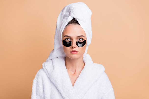 Фото серьезной молодой женщины носить глазной пластырь банный халат изолирован на пастельно-бежевом фоне - Фото, изображение