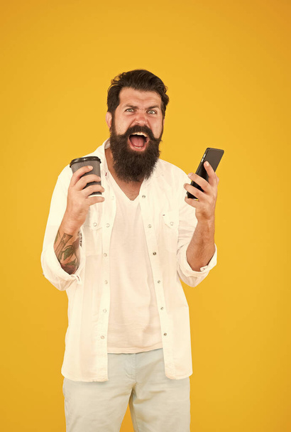 shouting guy with beard and moustache has mobile phone. morning routine. use modern technology. angry bearded man holding phone while drink coffee. brutal hipster drinking takeaway beverage - Zdjęcie, obraz