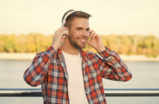 улыбающийся бородатый мужчина слушать музыку в цифровых наушниках носить клетчатую рубашку на открытом воздухе, технологии - Фото, изображение