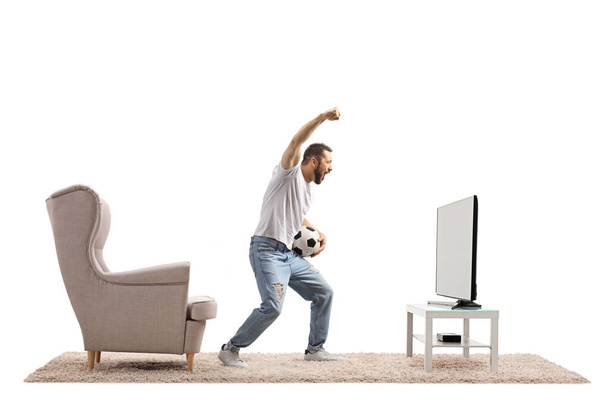 Young man in front of a tv holding a soccer ball and cheering with hand up isolated on white background - Fotó, kép