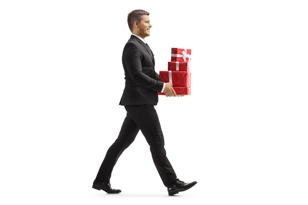 Full length profile shot of a man in black suit walking and carrying a pile of presents isolated on white background - Foto, Imagem