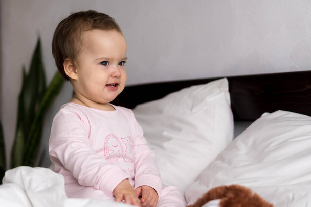 Authentic portrait caucasian little infant chubby baby girl or boy in pink sleepy upon waking looking at camera smiling cute and grimaces in white bed. Child care, Childhood, Parenthood, life concept - Φωτογραφία, εικόνα