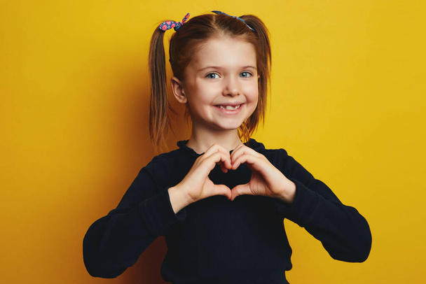 Pretty young cute girl makes heart gesture, isolated over yellow background - Foto, immagini