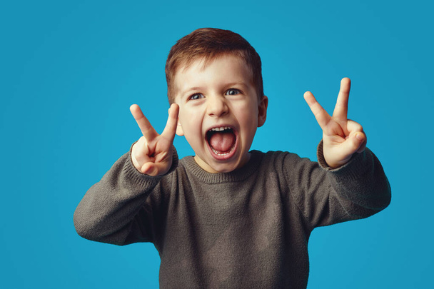 Χαριτωμένο παιδί σηκώνει τα χέρια δείχνει την ειρήνη χειρονομία με ανοιχτό το στόμα, γελάει ευτυχισμένος - Φωτογραφία, εικόνα