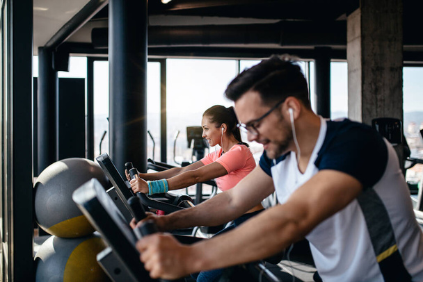 Young and attractive woman and man biking in the fitness gym. They exercising legs and doing cardio workout while riding cycling machines. - Zdjęcie, obraz