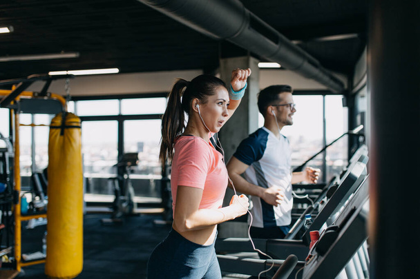Young fit man and woman running on treadmill in modern fitness gym. - Zdjęcie, obraz