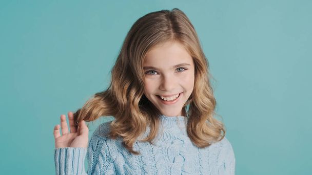 Charmant blond tiener meisje met golvend haar op zoek schattig glimlachen op camera geïsoleerd op blauwe achtergrond - Foto, afbeelding