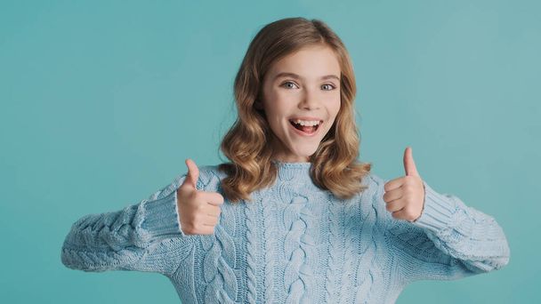 Cheerful blond teenage girl keeping thumbs up on camera and smiling isolated on blue background. Body language concept - Fotó, kép