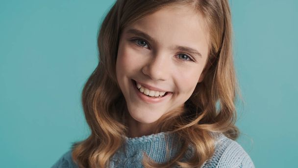 Charmantes blondes Teenager-Mädchen mit welligem Haar, das vor blauem Hintergrund in die Kamera lächelt. Glückliche Miene - Foto, Bild