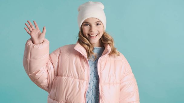 Pretty blond teenage girl dressed in pink down jacket and hat greeting friend over blue background. Charming girl waving hello - 写真・画像
