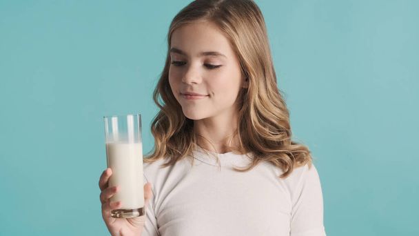 Attractive blond teenager girl holding glass of milk posing on camera over on blue background - Foto, imagen
