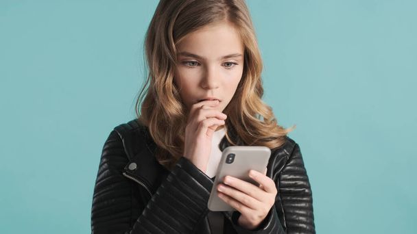 Portrait of blond teenager girl getting message from friend on smartphone looking thoughtful over colorful background. Deep in thoughts - Foto, imagen
